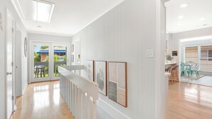 A hallway with a wooden floor, white walls, and a white railing. There are framed artworks on the wall and a view of a balcony with outdoor furniture. - The Big Tybreezy (Tybee Island)
