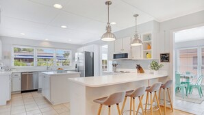 A modern kitchen with white cabinetry, a central island with bar stools, stainless steel appliances, and a view to an outdoor patio area. - The Big Tybreezy (Tybee Island)