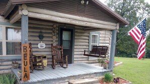 A log cabin with a porch, a rocking chair, a bench, and a welcome sign. - Beula 1869 cabin in "The Land" (Calico Rock)