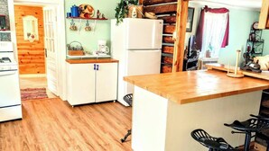 A compact kitchen with wooden flooring, a white refrigerator, a wooden counter, and a small dining area with a chair. - Beula 1869 cabin in "The Land" (Calico Rock)