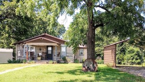A log cabin with a front porch, a small shed, and a well-maintained lawn. - Beula 1869 cabin in "The Land" (Calico Rock)