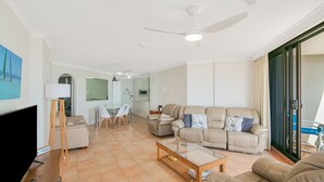 Un salon spacieux avec un canapé, une table basse et un téléviseur. On y trouve également un coin repas avec des chaises et une table, ainsi qu’une cuisine à l’arrière-plan.