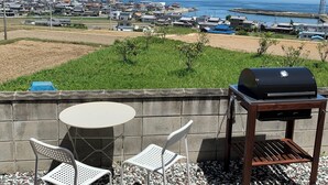 Eine Terrasse mit rundem Tisch und zwei Stühlen, einem Grill und Blick auf eine Küstenstadt.
