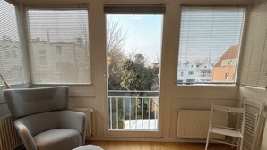 A spacious room with a grey sofa, a small table, and a radiator. There are large windows with blinds, and a balcony is visible through the door.
