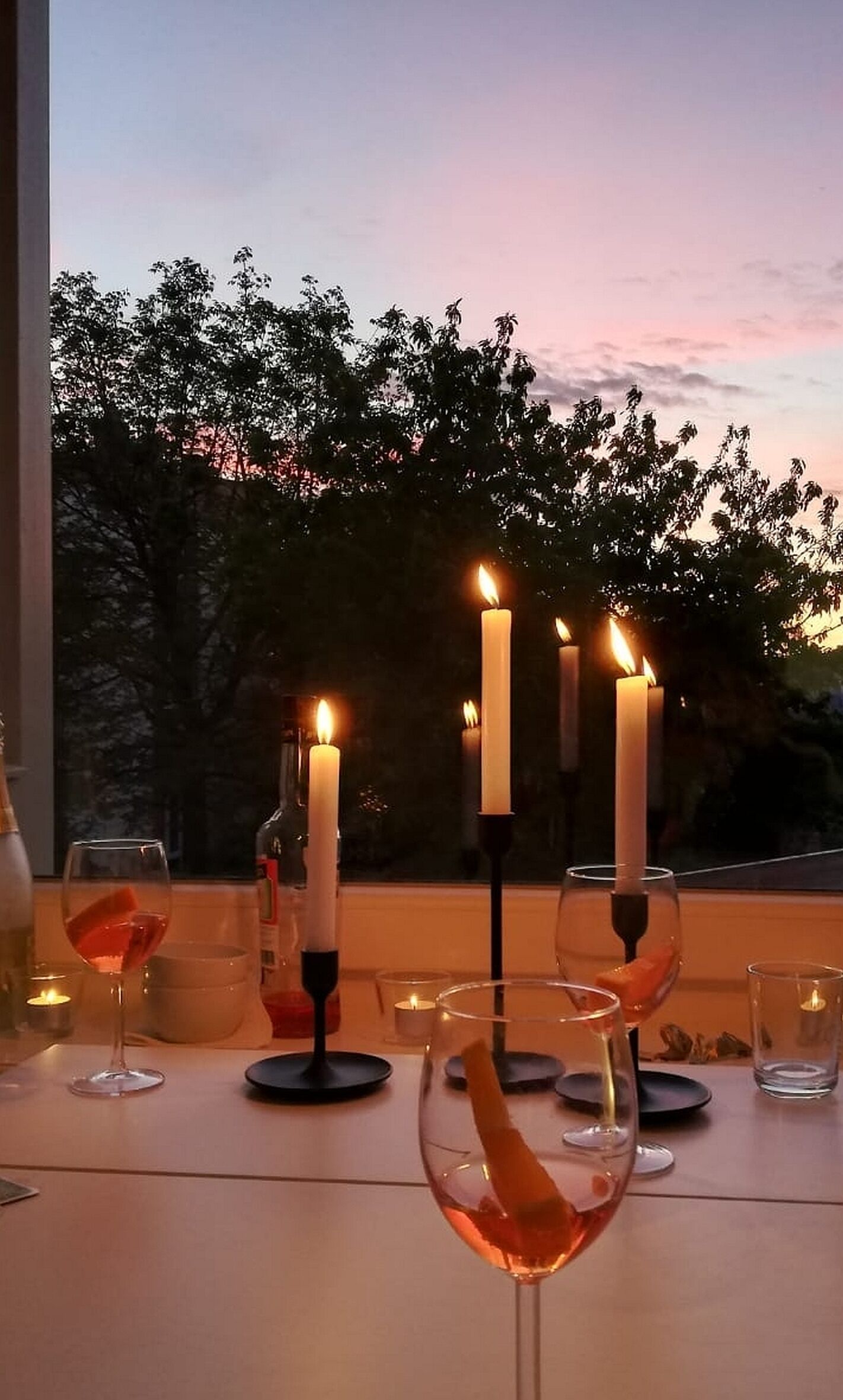 A table set for dining with candles, wine glasses, and a view of trees and sky at dusk.