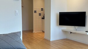 A modern living room with a flat-screen TV, a grey sofa, wooden flooring, and a kitchen visible through an archway.