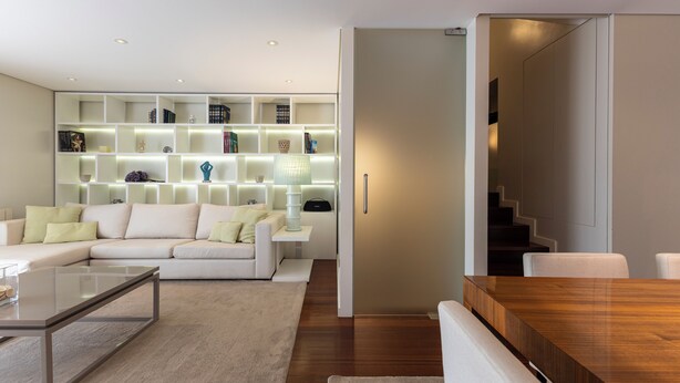 Un salon moderne avec un canapé blanc, une table basse en verre et une table à manger en bois. On y trouve une bibliothèque avec des étagères éclairées et un escalier sur le côté droit.