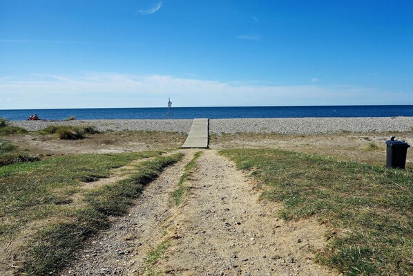 Un sendero de grava que lleva a una playa con un paseo marítimo de madera, un bote de basura y una torre de salvavidas a lo lejos.