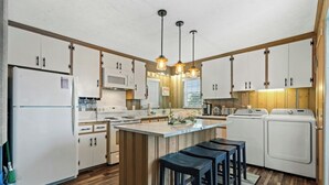 A modern kitchen with white cabinets, a central island with stools, a microwave, stove, and washing machines. - Channeling The Sun-4BR/2BA Channel House- Pets (North Myrtle Beach)