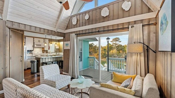 A spacious living area with a ceiling fan, a kitchen in the background, and a balcony with a view of trees. - Channeling The Sun-4BR/2BA Channel House- Pets (North Myrtle Beach)