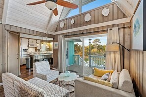 A spacious living area with a ceiling fan, a kitchen in the background, and a balcony with a view of trees. - Channeling The Sun-4BR/2BA Channel House- Pets (North Myrtle Beach)