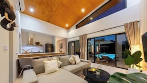 A modern living room with a sofa, a coffee table, and a view of a swimming pool through large glass doors. - Sereniwood 2 - 3BR Villa with Private Pool (Koh Samui)