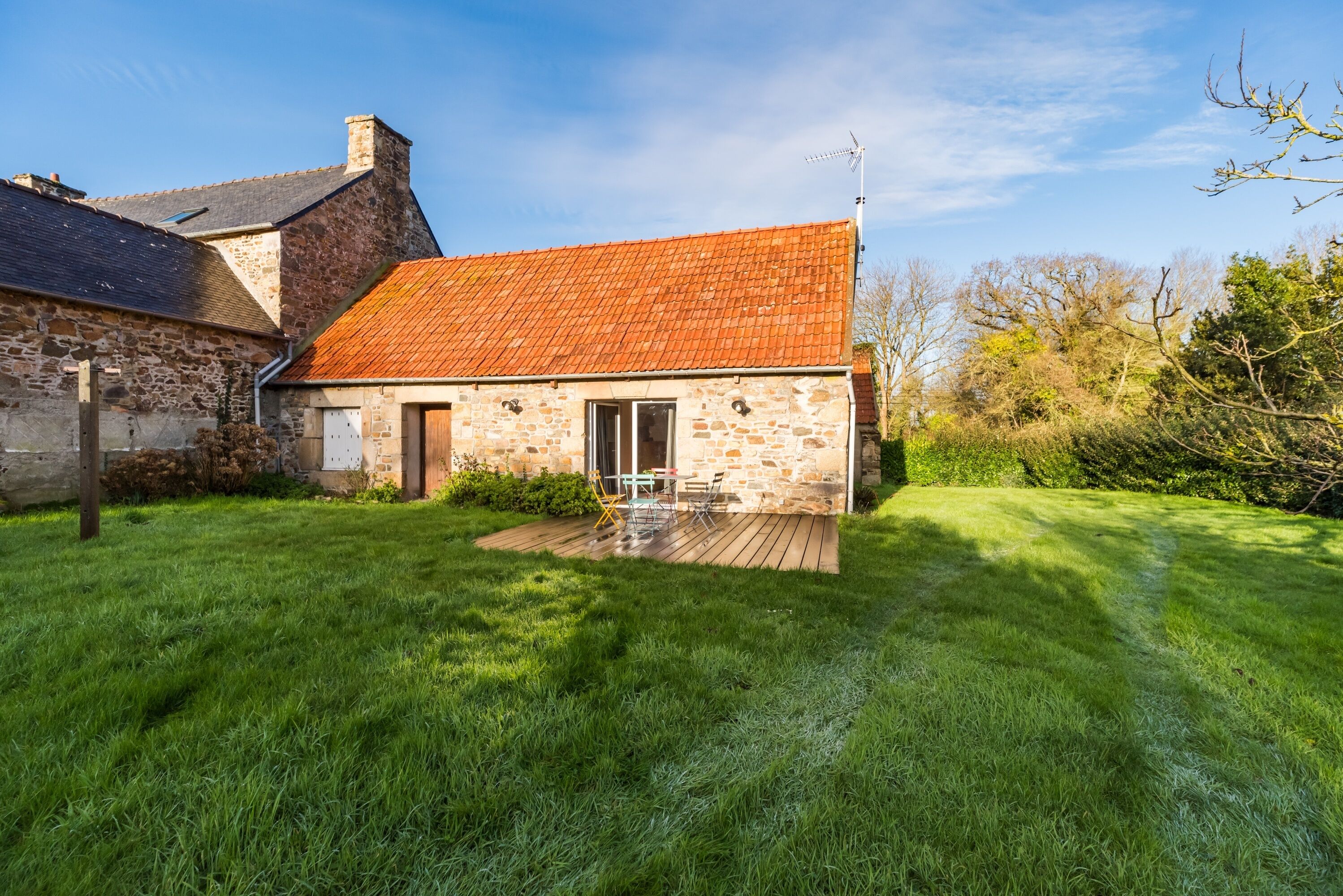 A stone house with a red-tiled roof, a wooden deck, and a grassy yard.
