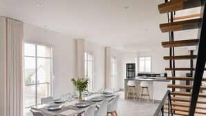 Ein moderner Essbereich mit einem langen Tisch, gedeckt für sechs Personen, einer Küche im Hintergrund und einer Treppe, die zu einer oberen Ebene führt.