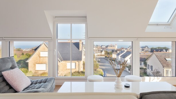 Ein modernes Wohnzimmer mit einem grauen Sofa, einem weißen Couchtisch und Blick auf eine Wohngegend durch große Fenster.