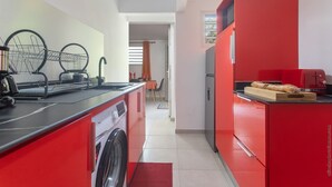 A compact kitchen with red cabinetry, a washing machine, and a dining area in the background.
