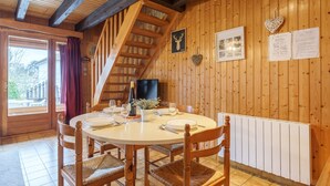 A wooden interior with a round dining table, chairs, a staircase, and a view through a door.