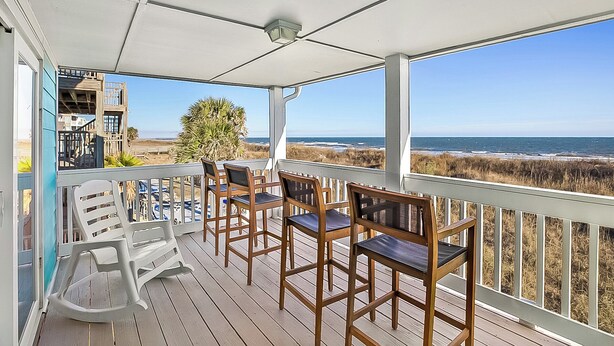 A covered outdoor deck with wooden chairs and a white rocking chair, overlooking a beach with dunes and the ocean. - Twin Palms Beach House (Sleeps up to 26) (North Myrtle Beach)