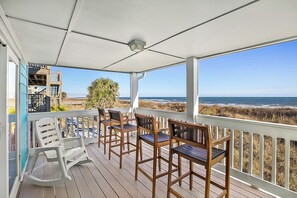 En takförsedd uteplats med trämöbler och en vit gungstol, med utsikt över en strand med sanddyner och havet.