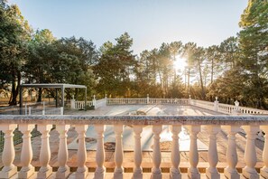 Une cour dallée de pierres, bordée d’une balustrade blanche, entourée d’arbres et baignée d’un ciel dégagé.