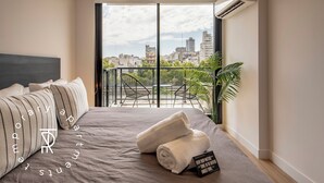 A modern hotel room with a large bed, a view of the cityscape, and a balcony with chairs and plants. - Top Rentals Palermo Soho (Buenos Aires)