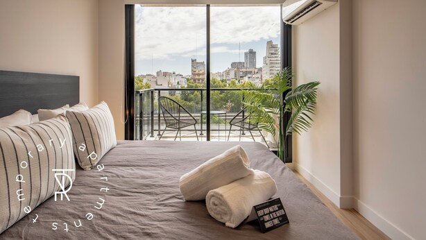 A modern hotel room with a large bed, a view of the cityscape, and a balcony with chairs and plants. - Top Rentals Palermo Soho (Buenos Aires)