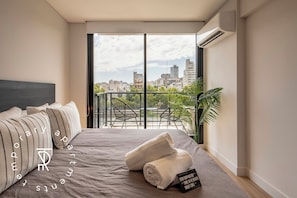 Une chambre d’hôtel moderne dotée d’un grand lit, offrant une vue sur le paysage urbain et comprenant un balcon avec des chaises et des plantes.