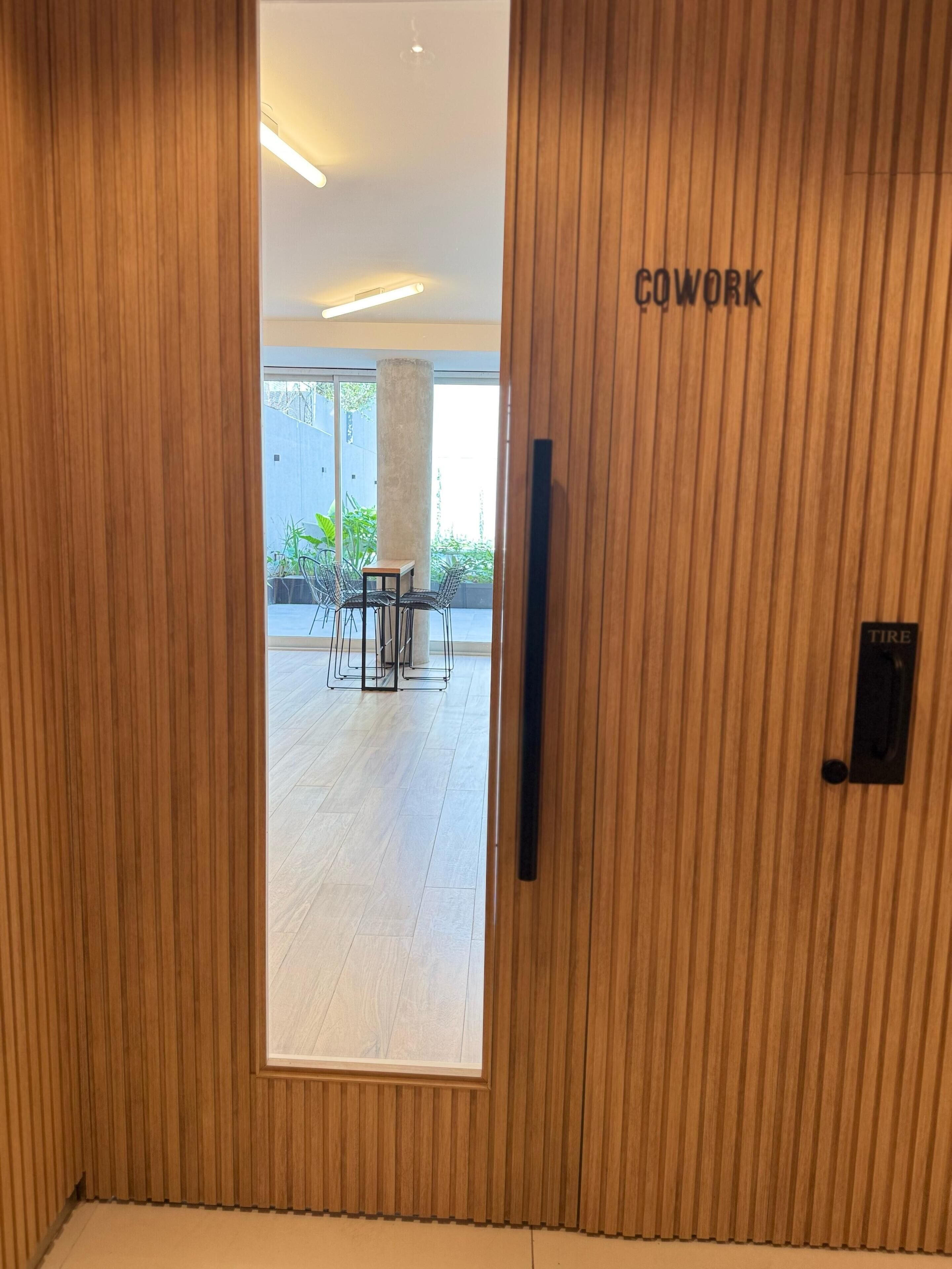 Een houten deur met de tekst 'COWORK', die leidt naar een ruimte met een groot raam, een plant en een tafel met stoelen.