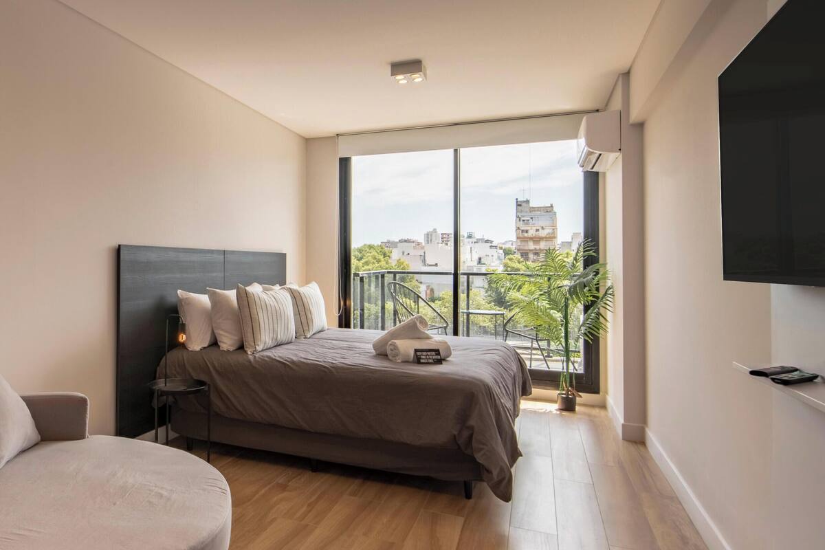 A modern bedroom with a large bed, a sofa, a TV, and a balcony with a city view.