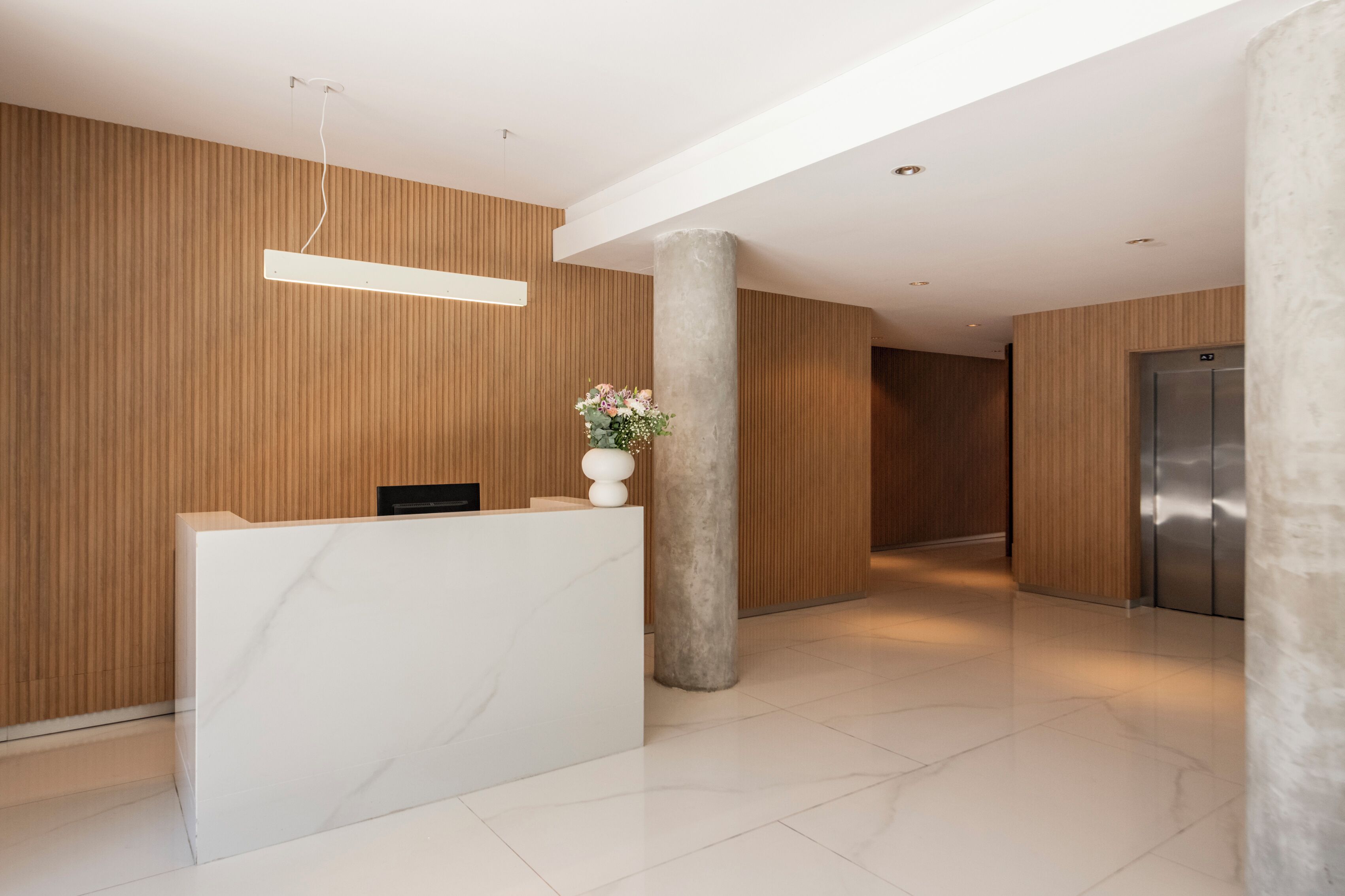 A modern reception area with a marble reception desk, wooden panel walls, a concrete pillar, and a stainless steel elevator.