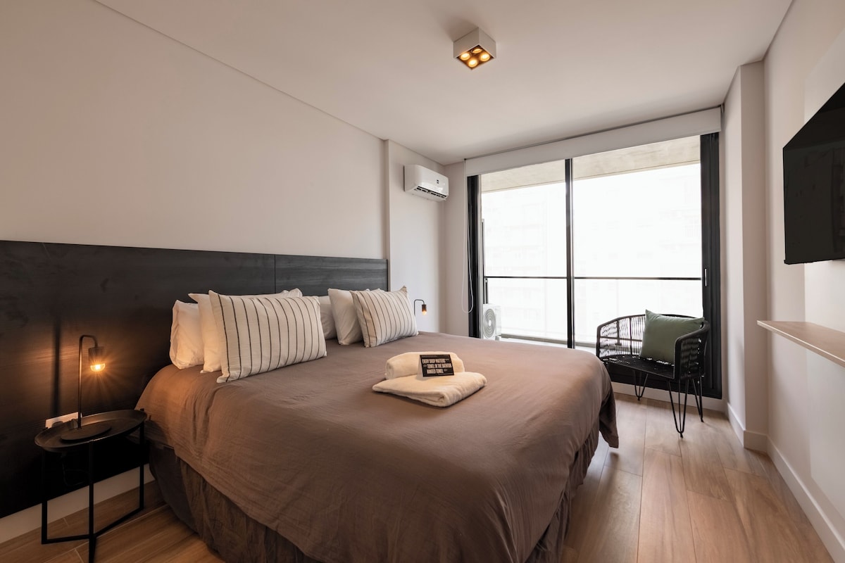 A modern bedroom with a large bed, bedside lamp, and a view of the outdoors through a large window.