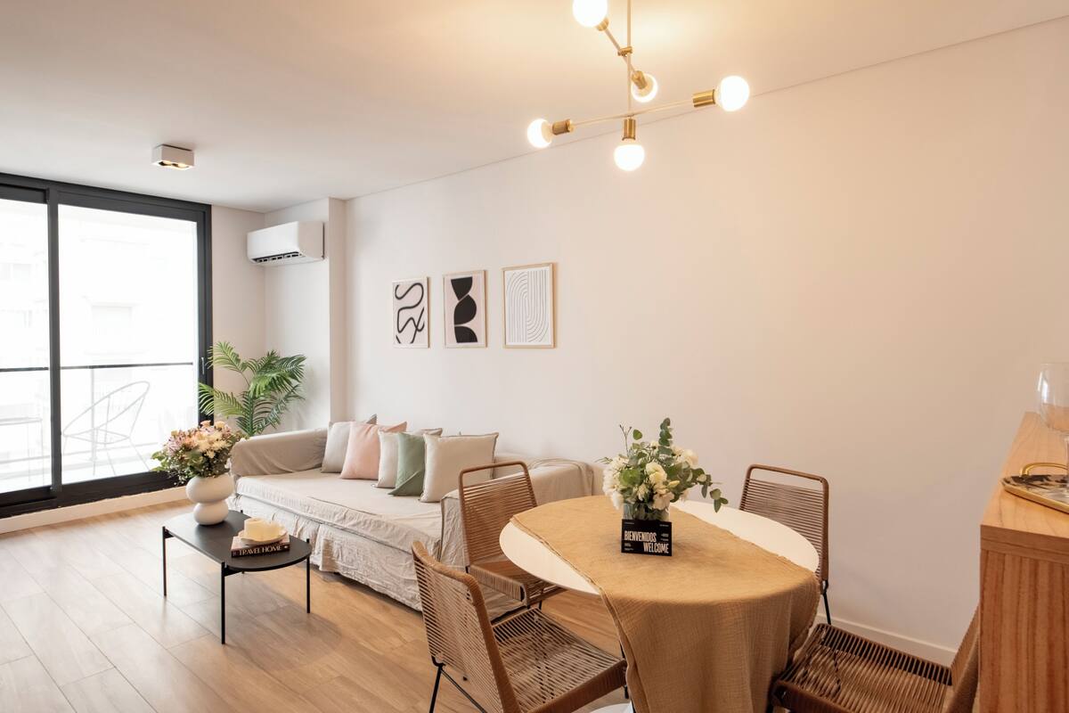 A modern living room with a sofa, dining table, and chairs. There is a large window providing natural light and a view outside.