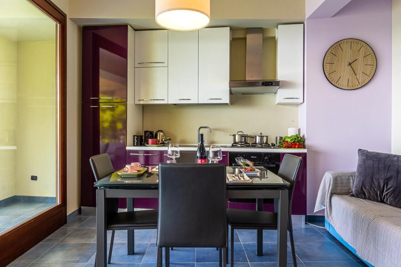 A compact kitchen with white cabinets, a dining table with chairs, a clock on the wall, and a sofa in the corner.