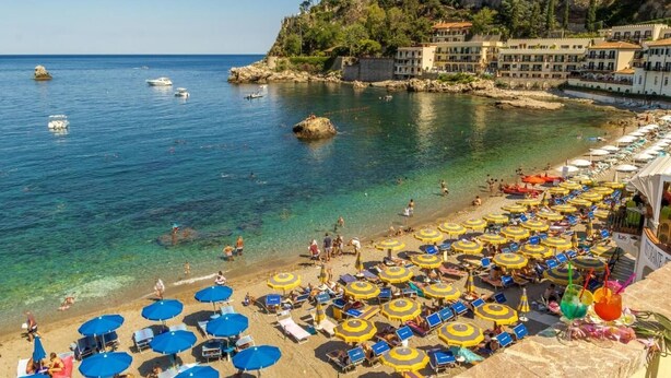 Une plage avec de nombreux parasols et des chaises longues, une côte rocheuse et des bâtiments en arrière-plan.