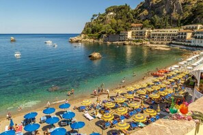 Una playa con numerosos sombrillas y sillas de playa, un litoral rocoso y edificios al fondo.