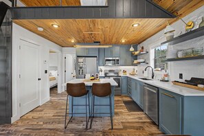A modern kitchen with wooden ceiling, blue cabinets, stainless steel appliances, and a central island with two chairs.