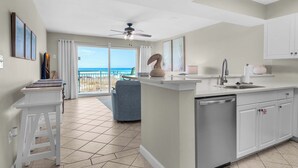 A modern kitchen with a central island, a dining area with a blue chair, and a view of the ocean through large windows.