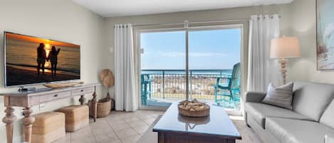A living room with a TV, sofa, and a balcony with a view of the ocean.