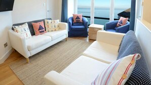 Una sala de estar con un sofá blanco, sillones azules y vista al mar a través de amplios ventanales.