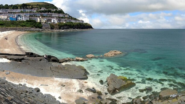 Un pueblo costero con playa despejada, costa rocosa y edificios en una colina.