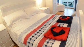 Una cama bien hecha con una colcha a rayas, una mesita de noche con lámpara y vista al mar a través de una ventana.