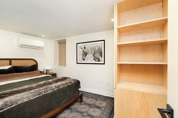 Un dormitorio con cama, cabecero, mesita de noche, lámpara, un cuadro en la pared y una estantería de madera.