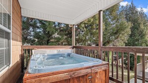 A wooden hot tub on a covered deck with a wooden railing, surrounded by trees. - BigFoot Inn, once you’ve seen it, you won’t want to leave! (PIGEON FORGE)