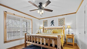 A bedroom with a wooden bed, a ceiling fan, a window with blinds, and two bedside tables with lamps. - BigFoot Inn, once you’ve seen it, you won’t want to leave! (PIGEON FORGE)