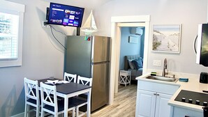 A compact kitchen with a built-in refrigerator, a dining table with chairs, a small island with a sink, and a flat-screen TV mounted above the refrigerator. - A beautiful tiny house in a beautiful little paradise for you to enjoy nature (Ocean Isle Beach)