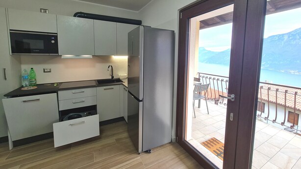 Cocina compacta con refrigerador, microondas y lavamanos. Una puerta corrediza de vidrio da a un balcón con vista a un lago y montañas.