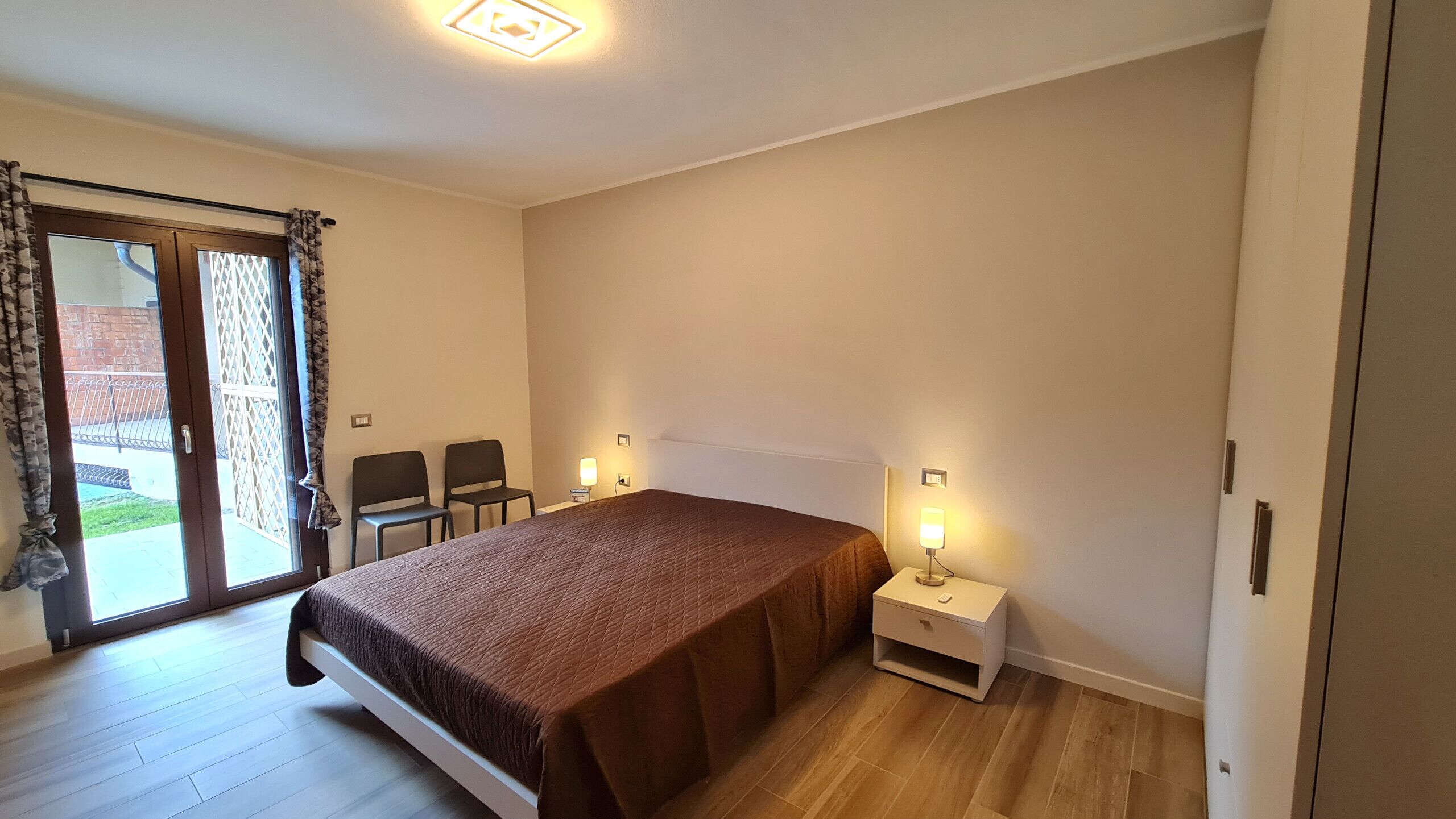 A bedroom with a bed, two chairs, a nightstand with a lamp, and a sliding glass door to the outside.