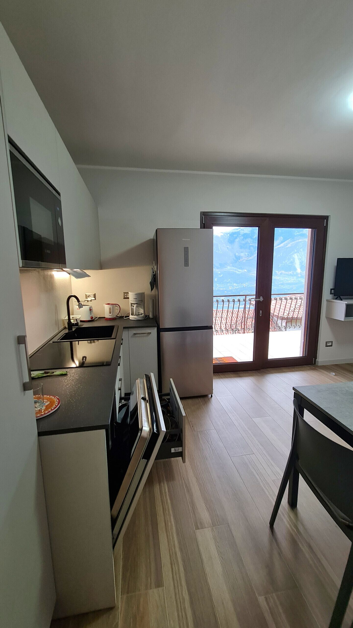 A compact kitchen with a refrigerator, stove, and microwave. There's a balcony with a view of the sky and a door leading outside.