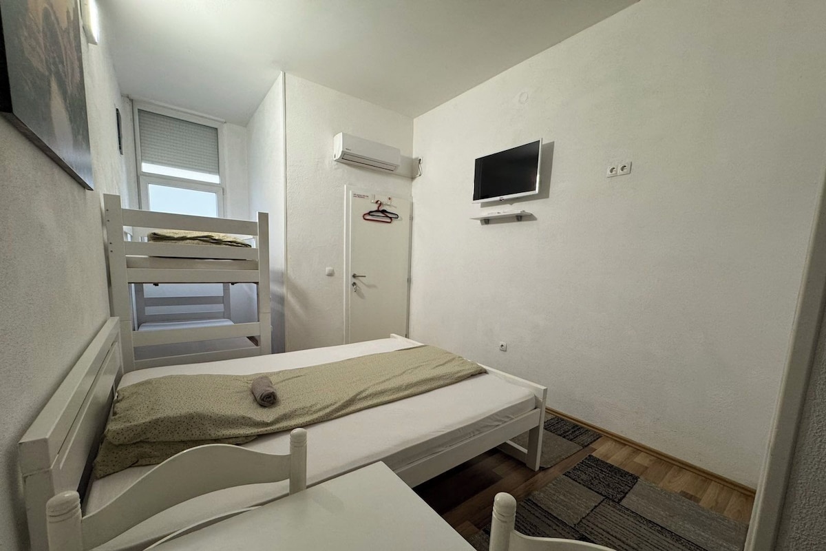A small, single-room accommodation with a bunk bed, a desk, a TV, and a door with a coat hanger.