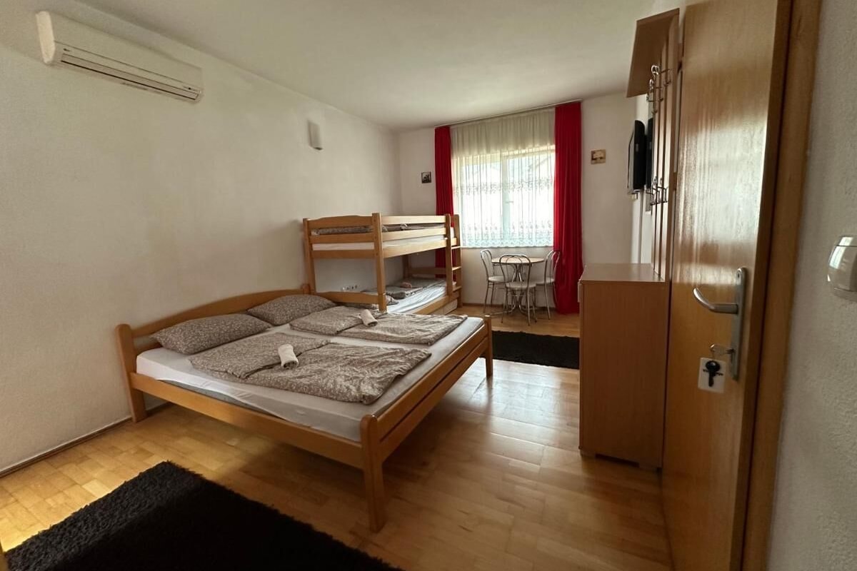 A single bed with a wooden frame and a mattress, a small table with chairs, a wardrobe, and a window with curtains.